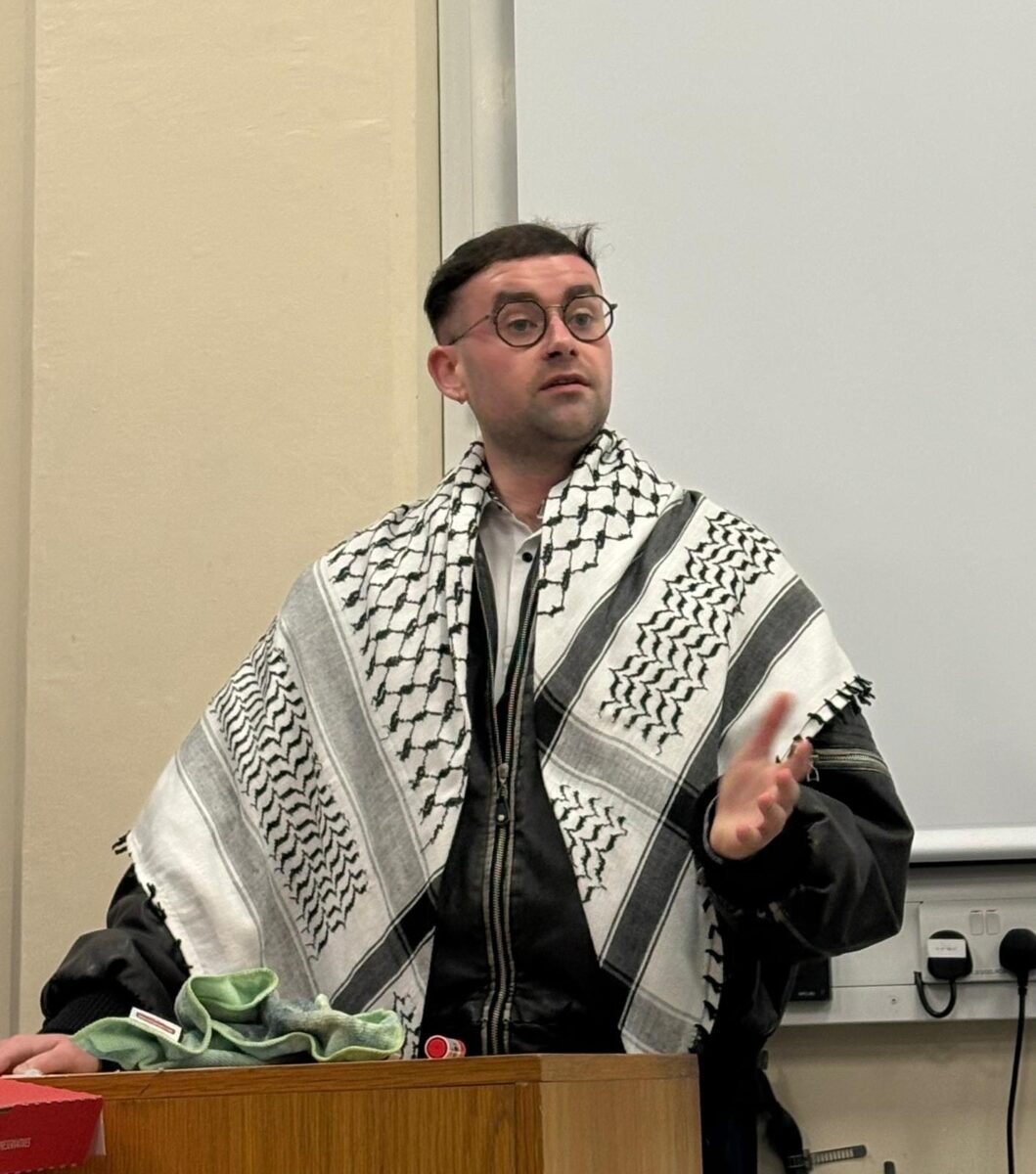 A young man speaks from a stand. he is wearing a Palestinian keffiyeh.